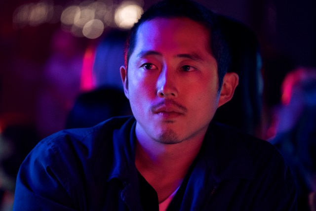 Steven Yuen as Danny Cho in Beef. He appears to be in a club. The club lighting is cool toned with purples, blues, and reds reflecting onto his face.